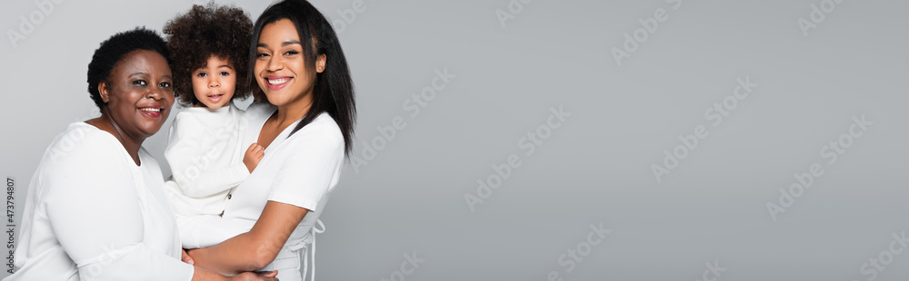 happy african american mature and young women with child looking at camera isolated on grey, banner happy african american mature and young women with child looking at camera isolated on grey, banner