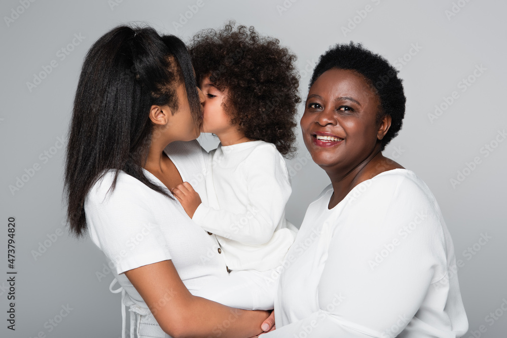 young african american woman kissing daughter near middle aged mom smiling at camera isolated on grey