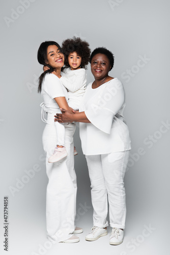 cheerful african american women in white clothes holding toddler girl on grey