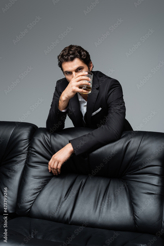 man in suit standing near black sofa and holding glass of whiskey isolated on grey man in suit standing near black sofa and holding glass of whiskey isolated on grey