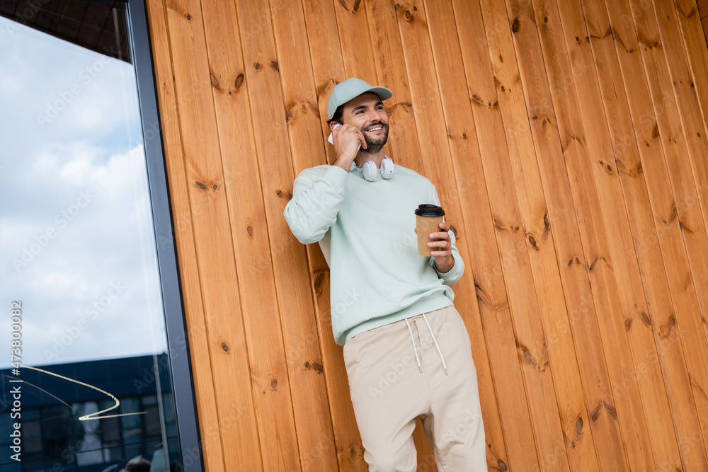 happy man in cap holding coffee to go while talking on smartphone near building happy man in cap holding coffee to go while talking on smartphone near building