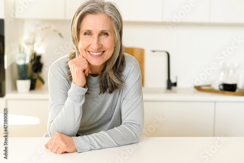 Elegant charming middle-aged woman with gray hair sitting at the countertop in bright modern kitchen and looks at the camera  smiling  put her chin on hands. Wellbeing concept