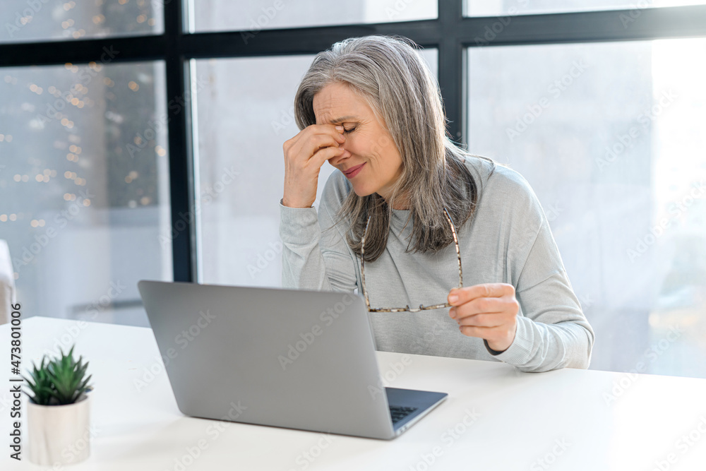 Tired mature female employee feel bored and tired, upset senior gray-haired woman sits with eyes closed in front of the laptop, leans head on the hand, feels headache and burnout Tired mature female employee feel bored and tired, upset senior gray-haired woman sits with eyes closed in front of the laptop, leans head on the hand, feels headache and burnout