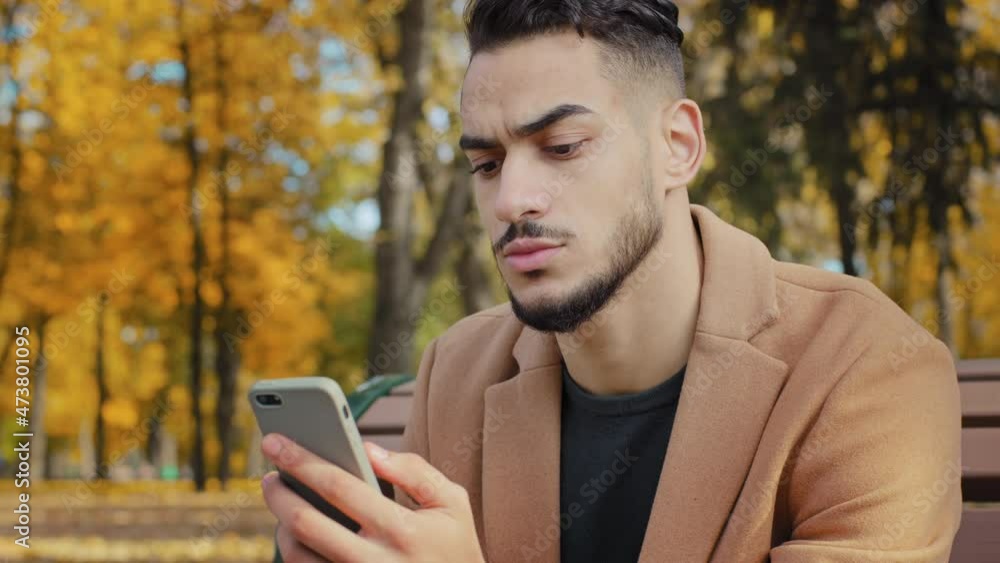 Portrait young serious guy use app hispanic man looking at phone screen browses news on telephone concentrated male sits on bench in autumn park read using device businessman holds smartphone outdoors Portrait young serious guy use app hispanic man looking at phone screen browses news on telephone concentrated male sits on bench in autumn park read using device businessman holds smartphone outdoors