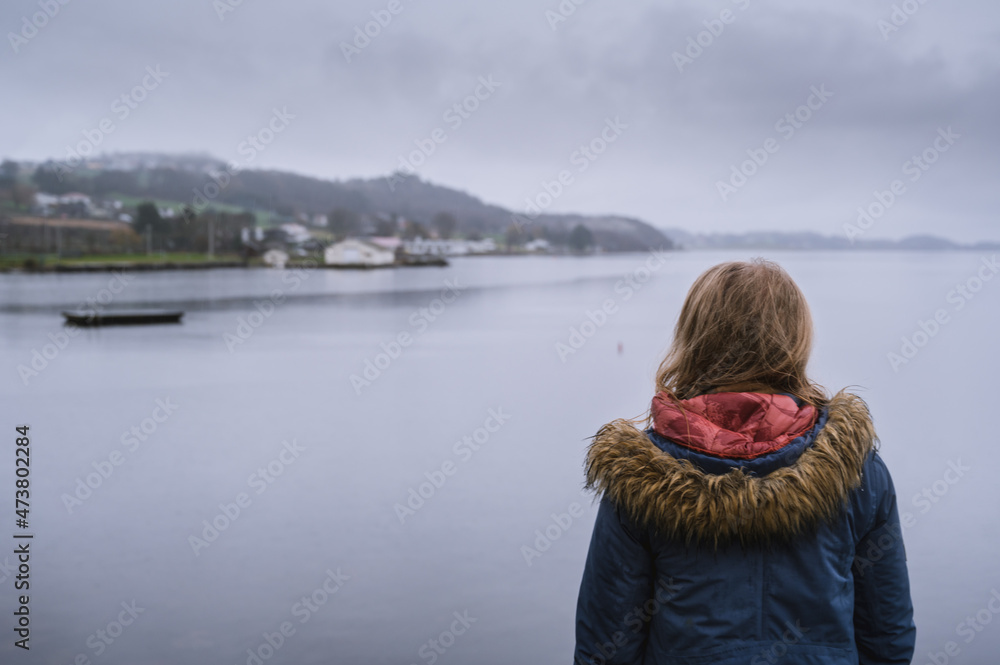 View of back of woman r in blue jacket near water. Cloudy rainy day. Hafrsfjord in Stavanger. View of back of woman r in blue jacket near water. Cloudy rainy day. Hafrsfjord in Stavanger.