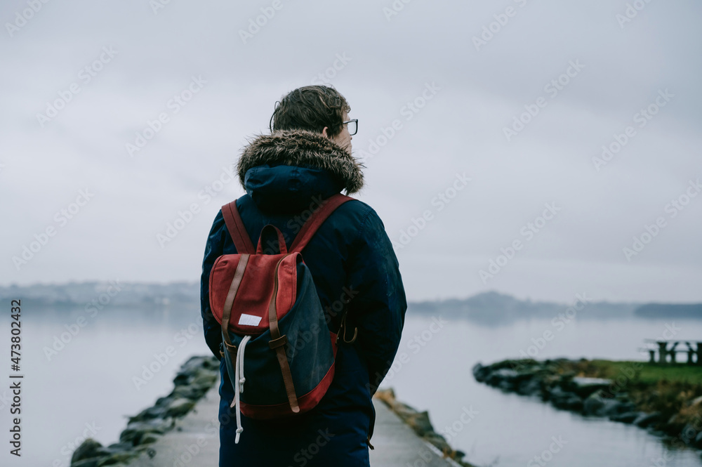 View of back of young man in blue coat with backpack on pier. Foggy fjord in background. Cloudy sky.