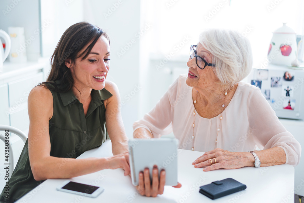 Senior woman talking with granddaughter using digital tablet while sitting at table Senior woman talking with granddaughter using digital tablet while sitting at table