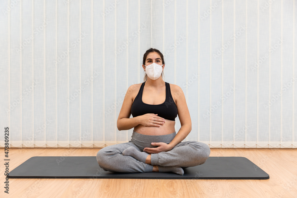 A healthy pregnant woman with face mask sitting on a mat doing yoga at home