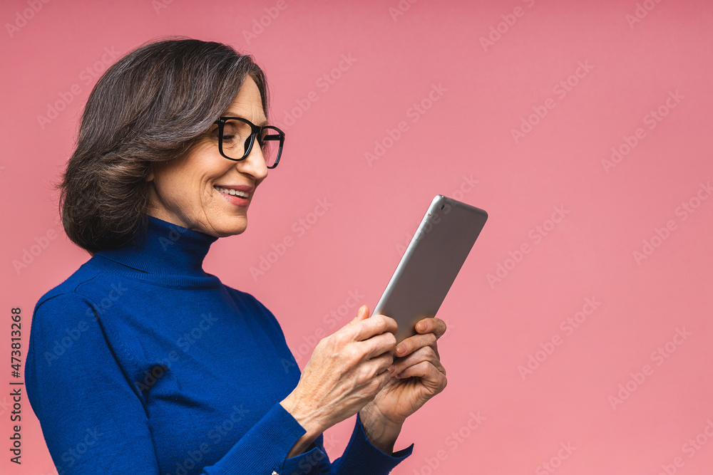 Portrait of smiling beautiful senior aged mature woman using tablet computer, isolated over pink background. Copy space for text.