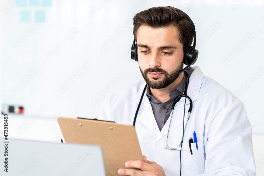 Handsome male doctor wearing headphone while reading report on clipboard in hospital Handsome male doctor wearing headphone while reading report on clipboard in hospital