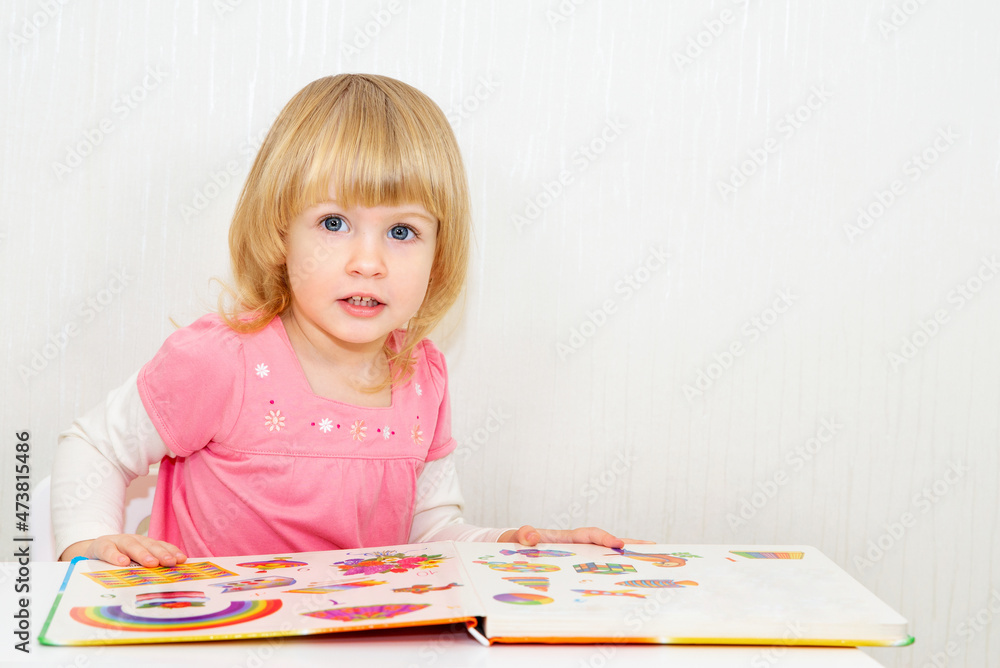 Cute little baby girl smiling. The child looks at the pictures in the book. Copy space. Light background. Cute little baby girl smiling. The child looks at the pictures in the book. Copy space. Light background.