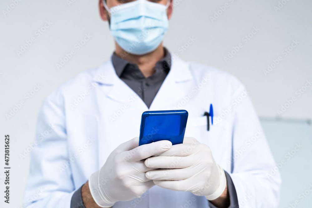 Male doctor text messaging through smart phone in hospital during COVID-19 Male doctor text messaging through smart phone in hospital during COVID-19