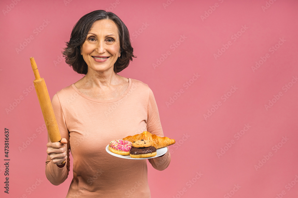Grandmother's bakery concept. Senior mature aged woman baker holding a rolling pin, donuts and croissant, isolated on pink background. Grandmother's bakery concept. Senior mature aged woman baker holding a rolling pin, donuts and croissant, isolated on pink background.