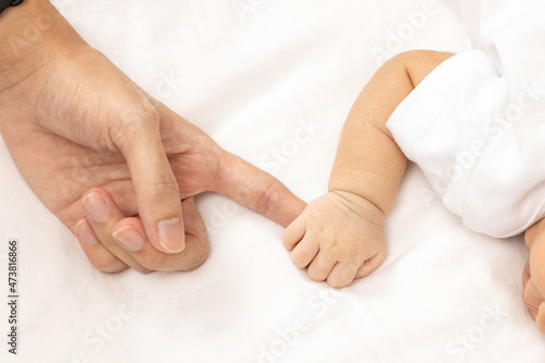 Little baby boy in grey pajamas holding mother's hand on the bed, close up