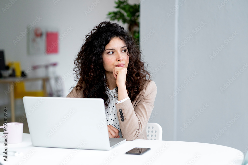 Businesswoman looking away while sitting with hand on chin at desk in office Businesswoman looking away while sitting with hand on chin at desk in office
