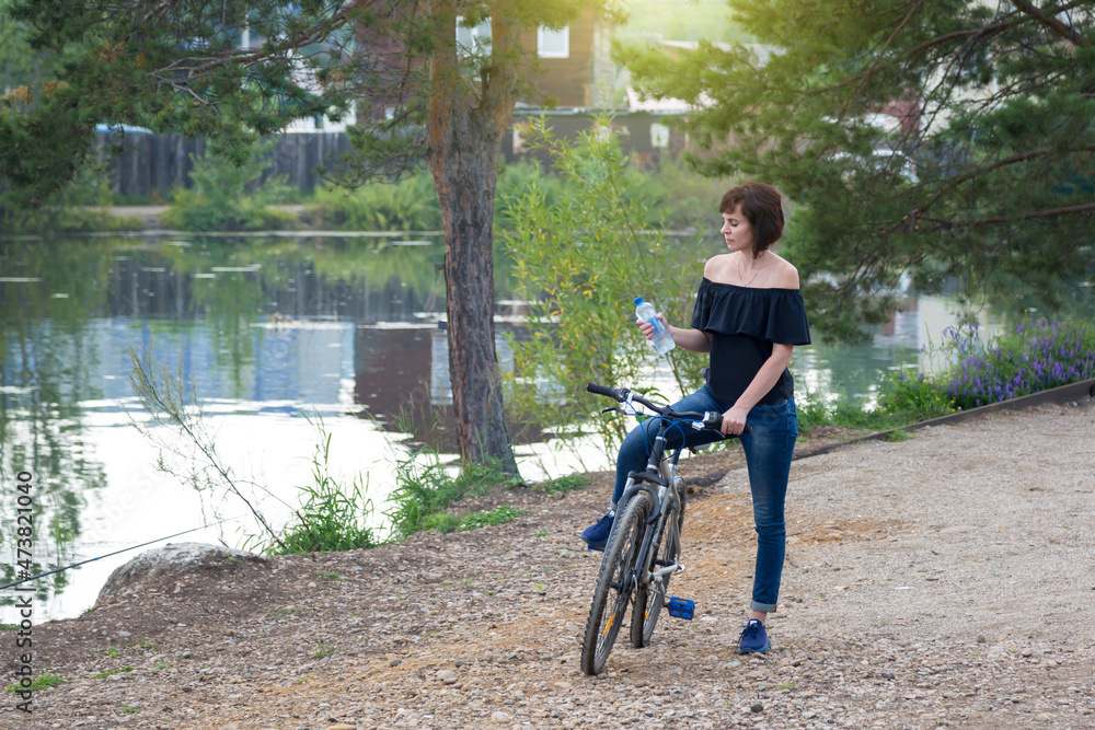 A brunette woman on a bicycle stopped by the river to drink water from a bottle.