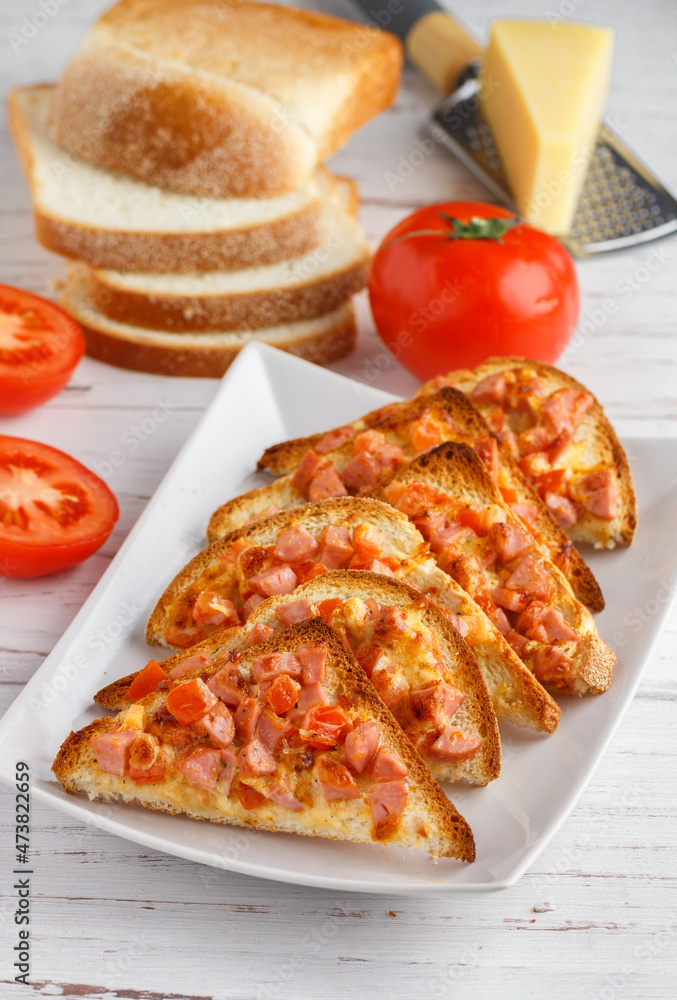 Homemade mini pizzas made of toast bread with ham or sausage, tomatoes, parmesan cheese and mozzarella and spices. Breakfast. White plate, wooden background. Brunch. Selective focus Homemade mini pizzas made of toast bread with ham or sausage, tomatoes, parmesan cheese and mozzarella and spices. Breakfast. White plate, wooden background. Brunch. Selective focus