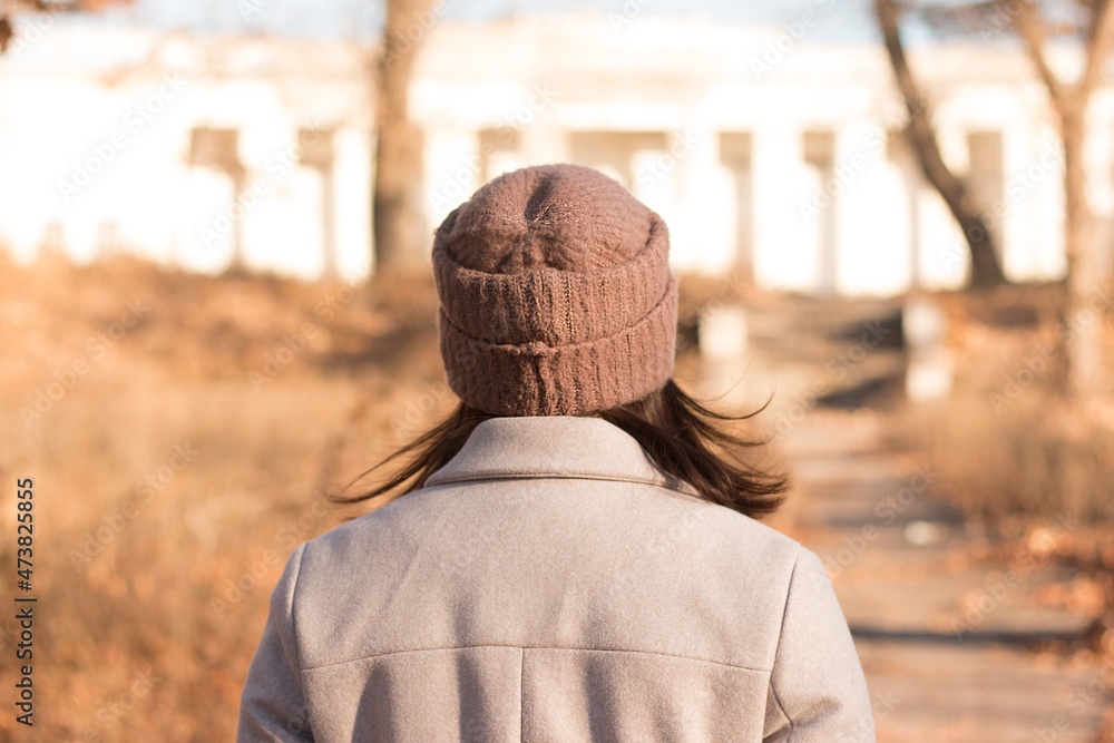 A girl with long dark hair in a coat and a knitted hat stands with her back to the camera against the background of an empty autumn street
