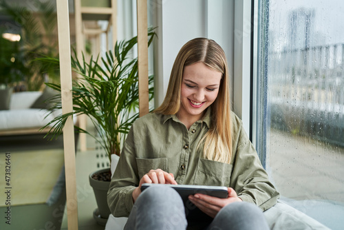 Smiling female intern using digital tablet while sitting by glass window in office photo