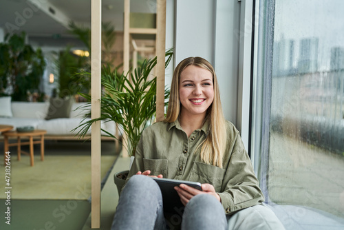 Smiling teenage female intern with digital tablet looking through window while sitting in office