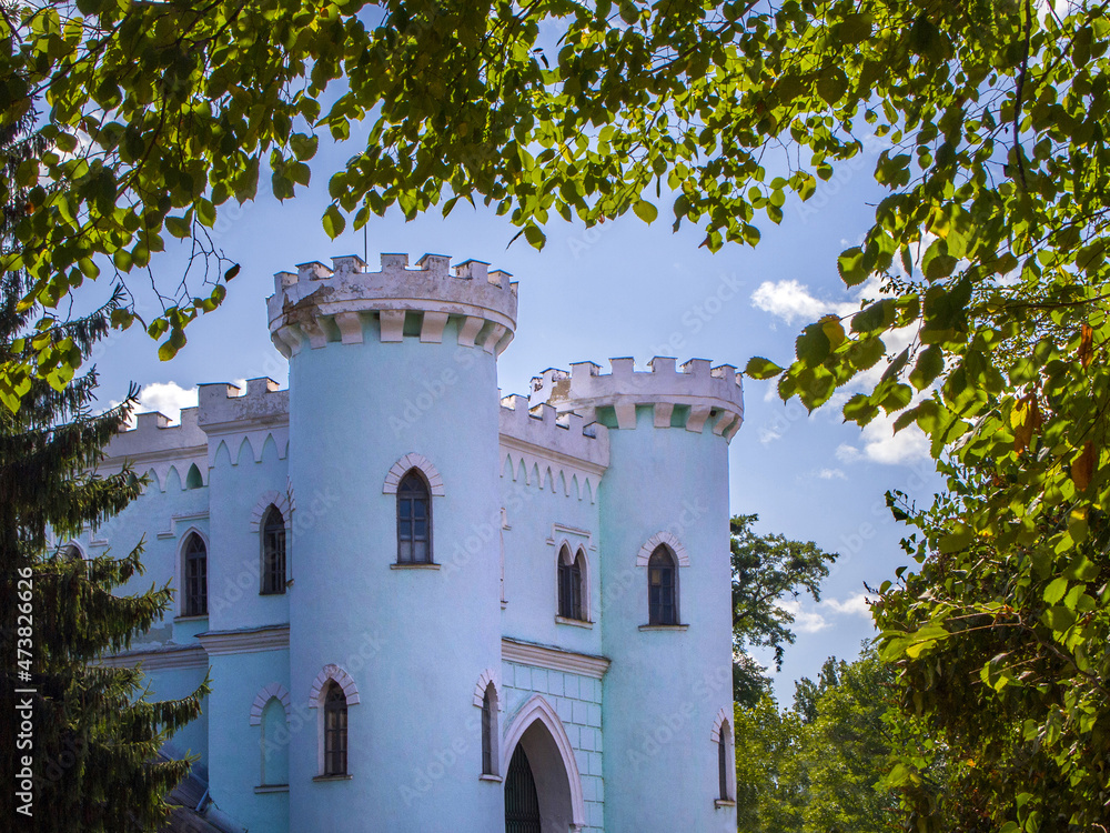 Fototapeta premium Part of a beautiful fairytale fortress surrounded by foliage and trees on a sunny day. The walls of the old building are painted in light blue and white. Magical atmosphere.