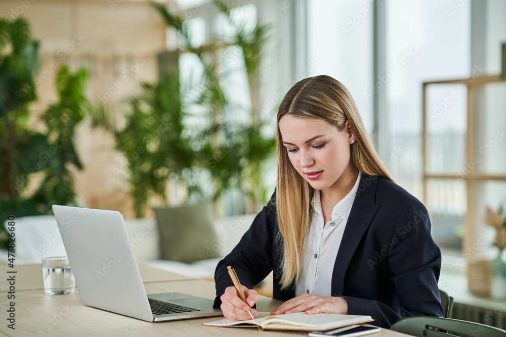 Female trainee writing in dairy while sitting at desk in office Female trainee writing in dairy while sitting at desk in office
