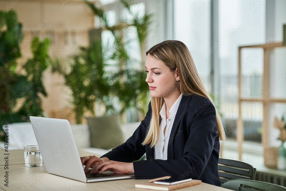 Blond female trainee typing on laptop while sitting at desk in office Blond female trainee typing on laptop while sitting at desk in office