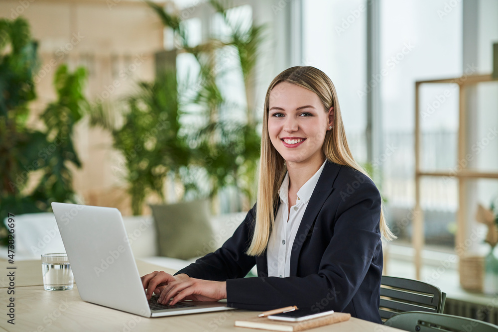 Smiling blond female trainee sitting with laptop at desk in office Smiling blond female trainee sitting with laptop at desk in office