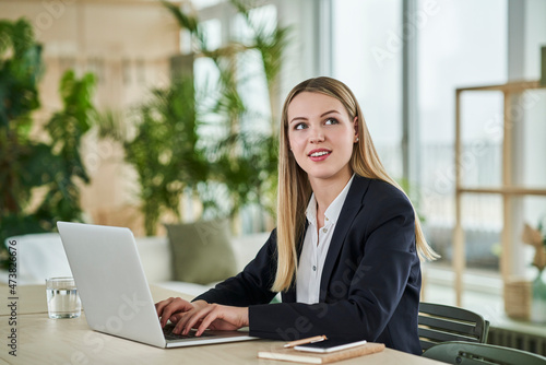 Blond female intern looking away while sitting with laptop at desk in office photo