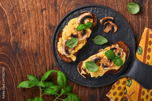 Scrambled eggs with fried mushrooms and basil on bread on black table background. Homemade breakfast or brunch meal - scrambled eggs and mushrooms sandwiches. Top view with copy space