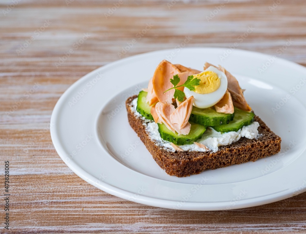 Smorrebrod, an open sandwich with boiled salmon, cream cheese, quail egg and cucumber slices on a white plate on a wooden background. Sandwich recipes. Smorrebrod, an open sandwich with boiled salmon, cream cheese, quail egg and cucumber slices on a white plate on a wooden background. Sandwich recipes.