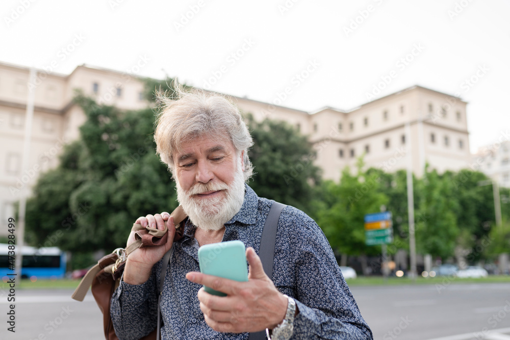 White bearded man carrying luggage while looking at smart phone in city White bearded man carrying luggage while looking at smart phone in city