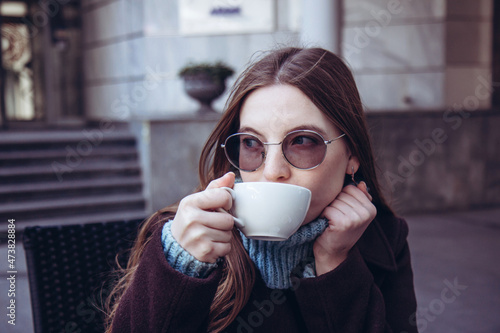 Beautiful woman with long hair in a dark coat sitting in a cafe on the street and drinks coffee. Lifestyle. Face close up