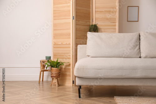 Stylish room with wooden folding screen and comfortable sofa near white wall. Interior design