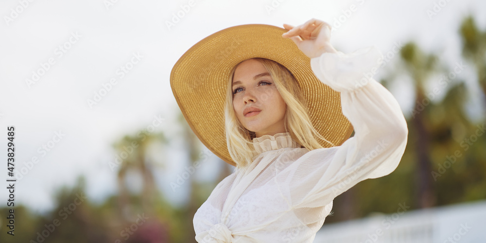 Stunning blonde woman in summer beach outfit relaxing outdoors against sea resort and palm trees on the background. A fashionable romantic young adult lady wearing a trendy vintage straw hat, white