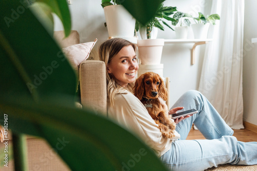 Smiling young woman with dog holding smart phone at home photo