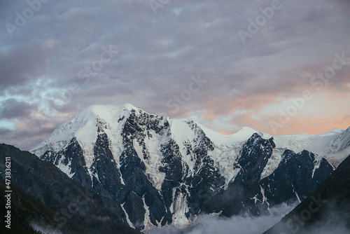 Atmospheric mountain landscape with great snowy mountains and low clouds in valley under violet orange dawn cloudy sky. Awesome alpine scenery with dense fog in mountain valley at sunset or at sunrise