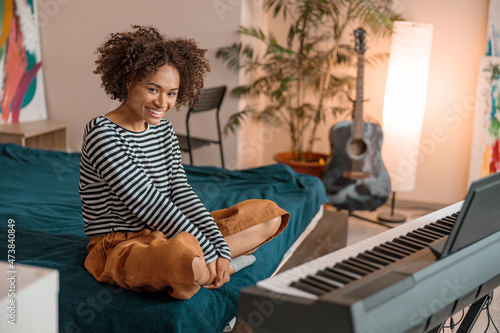 Cheerful multiracial woman looking at camera and smiling while sitting on bed in front of electronic musical instrument in bedroom photo