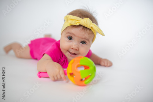cute, beautiful little girl in a pink bodysuit and a bandage lies on a white background, smiles and gnaws toys. beautiful baby. teeth are erupting. portrait of a baby. portrait of a beautiful girl