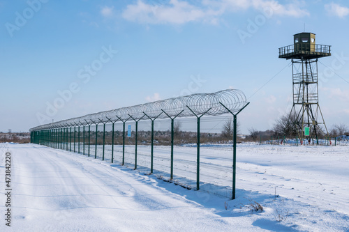 Border fence with barbed wire in front of the surveillance tower in a snow-covered field against a blue sky. Border protection and counteraction to illegal migration concept