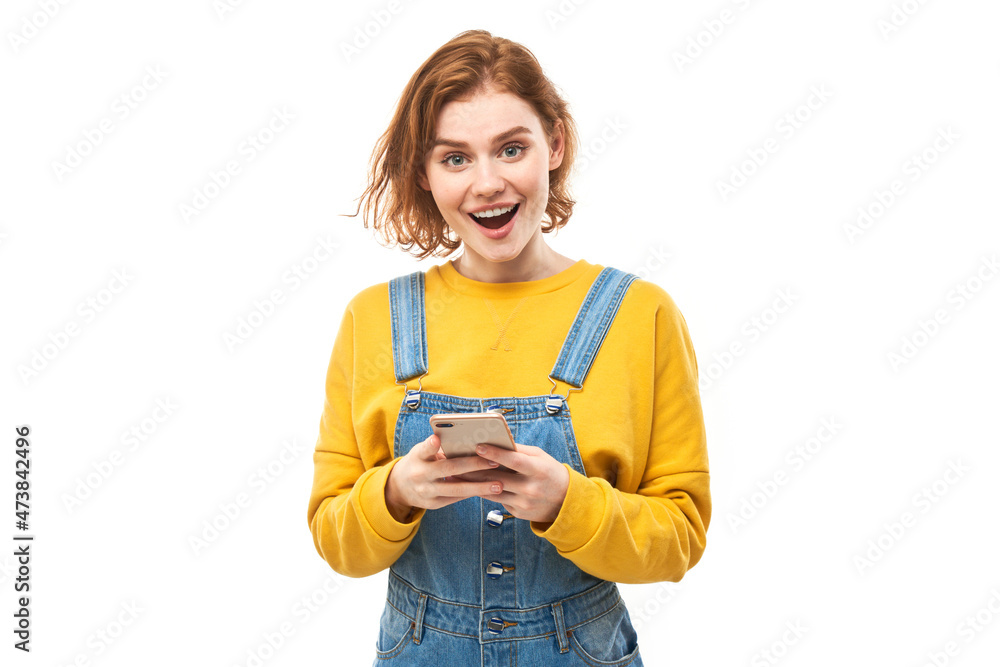 Happy smiling redhead girl holding smartphone in hands in yellow clothes isolated on white studio background, received joyful message Happy smiling redhead girl holding smartphone in hands in yellow clothes isolated on white studio background, received joyful message