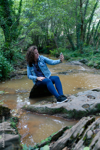 Vertical photo of wavy-haired Caucasian woman taking a selfie next to a stream.