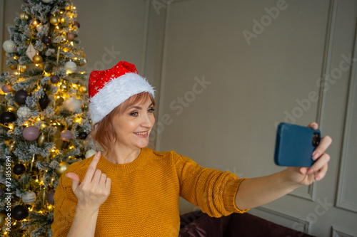 Happy woman in santa claus hat takes a selfie on their smart phone at christmas tree at home. Female are calling perents and friends through video call to congratulate them on new year. photo