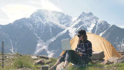 Wallpaper Mural Young woman in cap and sunglasses sits on hill top and types on laptop standing on rock against mountains and closes gadget closeup Torontodigital.ca