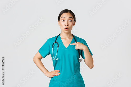 Covid-19, healthcare workers, pandemic concept. Surprised and ambushed asian female physician, nurse in scrubs pointing at herself, beaing named or chosen, standing white background photo