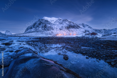 Sea coast with stones and beautiful snow covered mountain in winter at dusk. Beautiful fjord at night in Lofoten Islands, Norway. Nordic landscape with water, rocks, buildings, illumination, blue sky photo