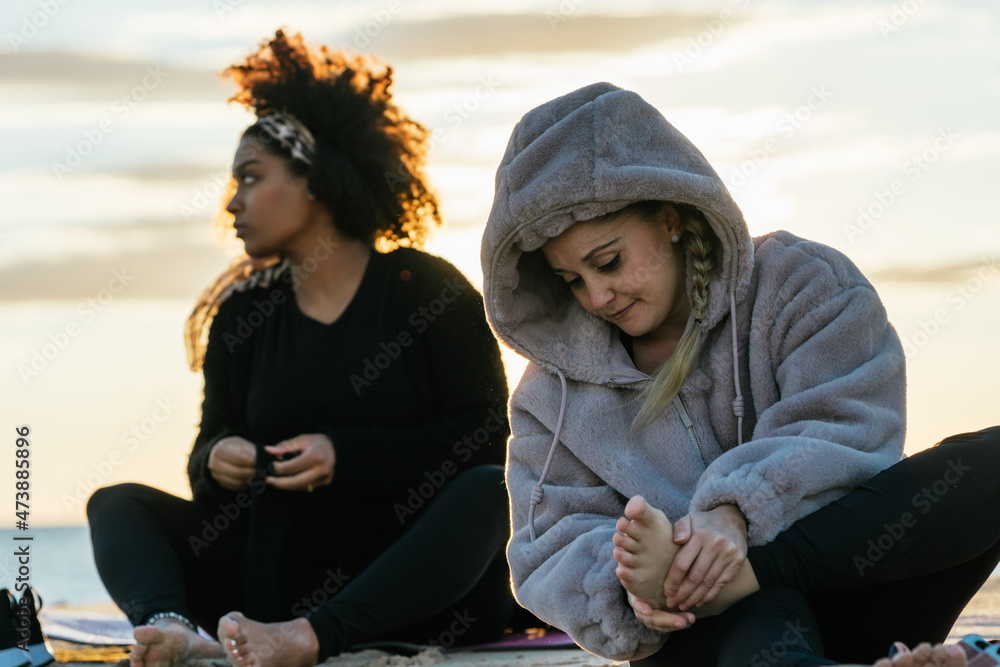 Two women in warm clothes sitting on a beach at sunset after a yoga class