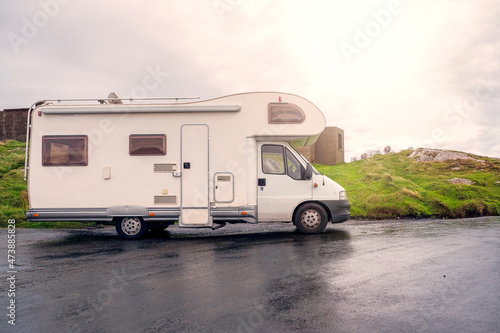 White motor home parked on wet asphalt car park. Sun flare. Travel and holiday in camper van. No people. Cloudy sky.