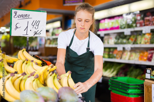 Obraz na plátně Fifteen-year-old girl who works part-time in a store as a trainee saleswoman puts recently brought bananas on the counter