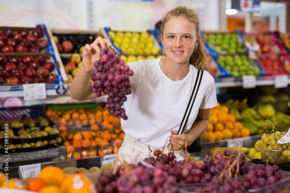 Portrait of a fifteen-year-old girl in a store at the fruit counter, chooses grapes, holding bunches in her hands